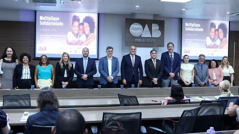 Foto de homens e mulheres em atrás de uma mesa. Dos lados esquerdo e direito há projeções com arte da campanha Multiplique Solidariedade.
