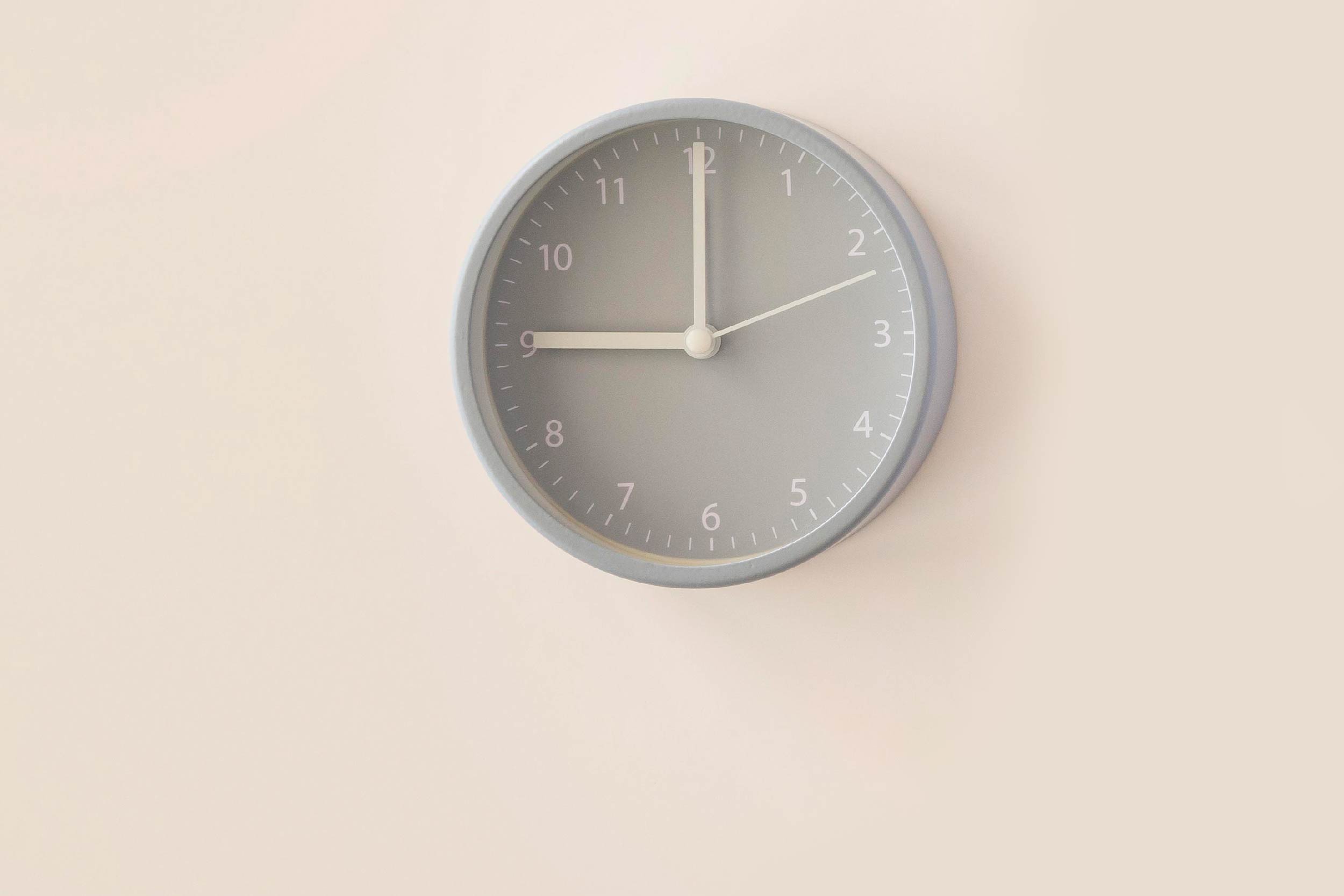 Foto de um relógio de parede cinza sobre fundo bege. O relógio marca 9 horas.
