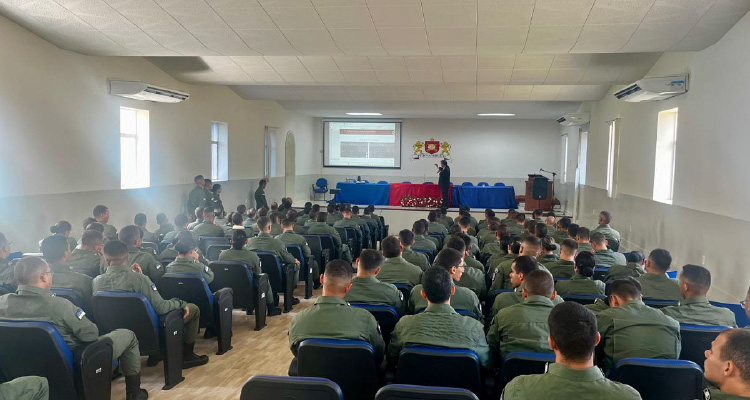 Foto mostra várias pessoas trajando farda da Polícia Militar sentadas em um auditório. No fundo da foto, perante uma mesa, um homem de terno fala ao público.