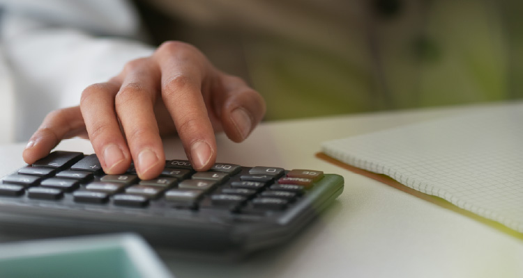 Imagem de mão digitando em calculadora junto a papéis espalhados
