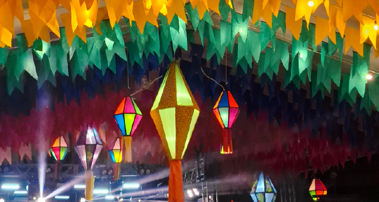 Imagem mostra bandeirolas e balões juninos pendurados no teto. No canto inferior esquerdo, há luzes de uma estrutura de palco.