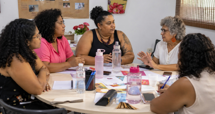 Cinco mulheres estão sentadas ao redor de uma mesa. No centro da mesa há papel, caneta e garrafas de água.