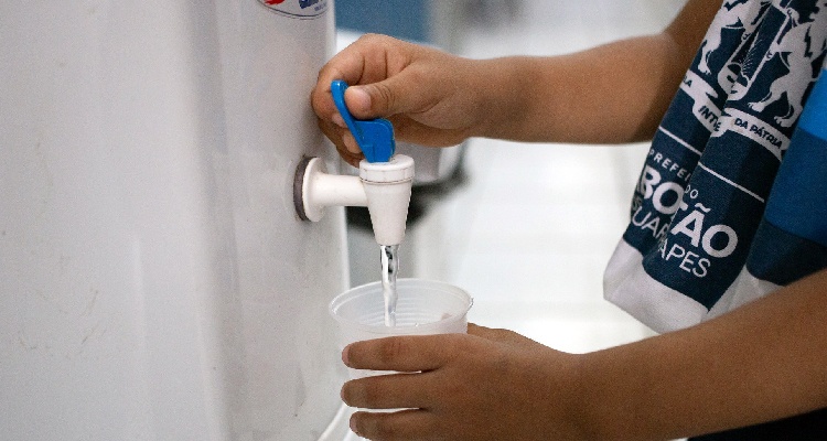 Imagem parcial de mãos infantis enchendo copo com água de uma torneira de bebedouro