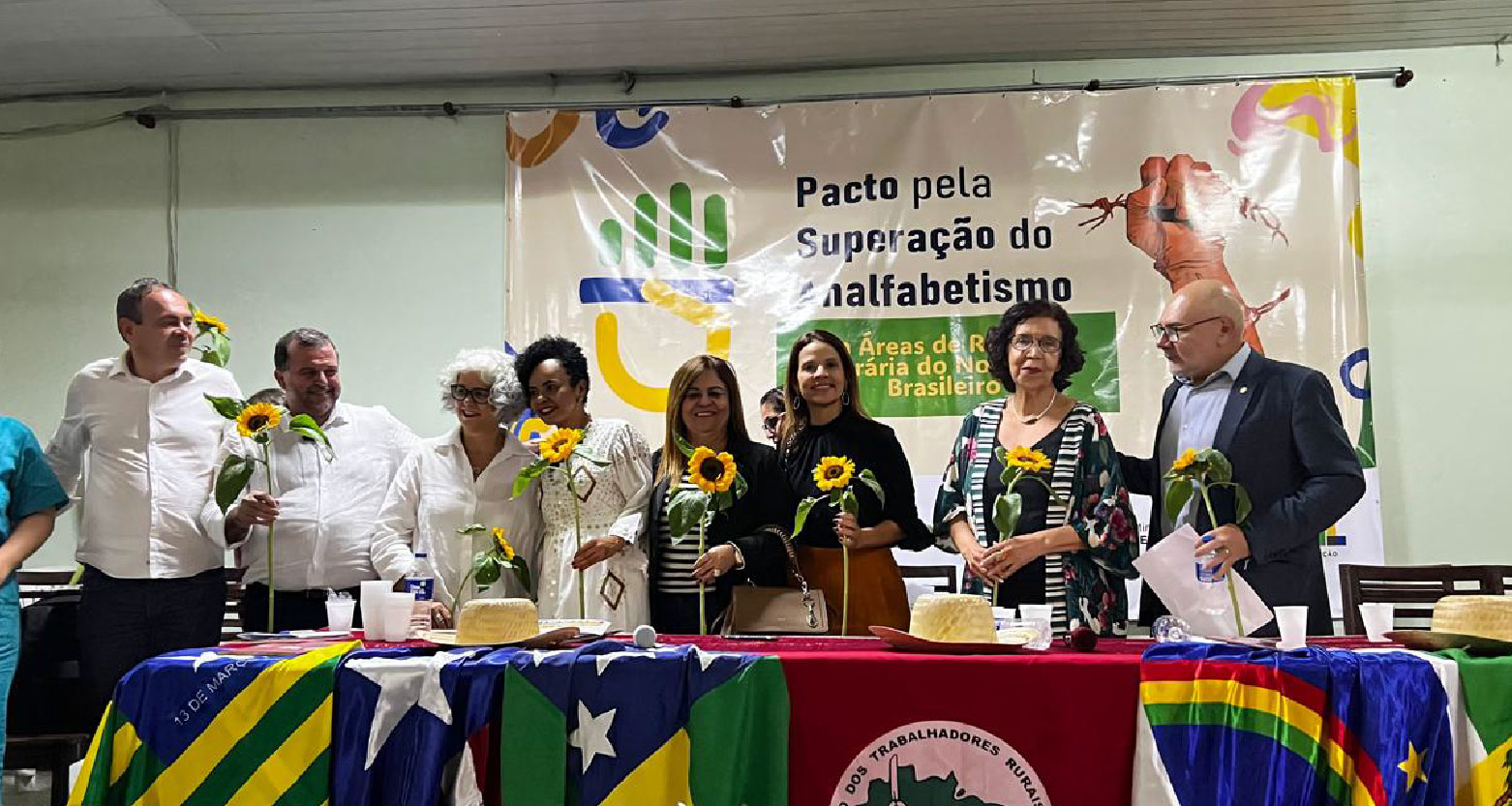 Fotografia de palestrantes do evento por trás da mesa principal, segurando girassóis