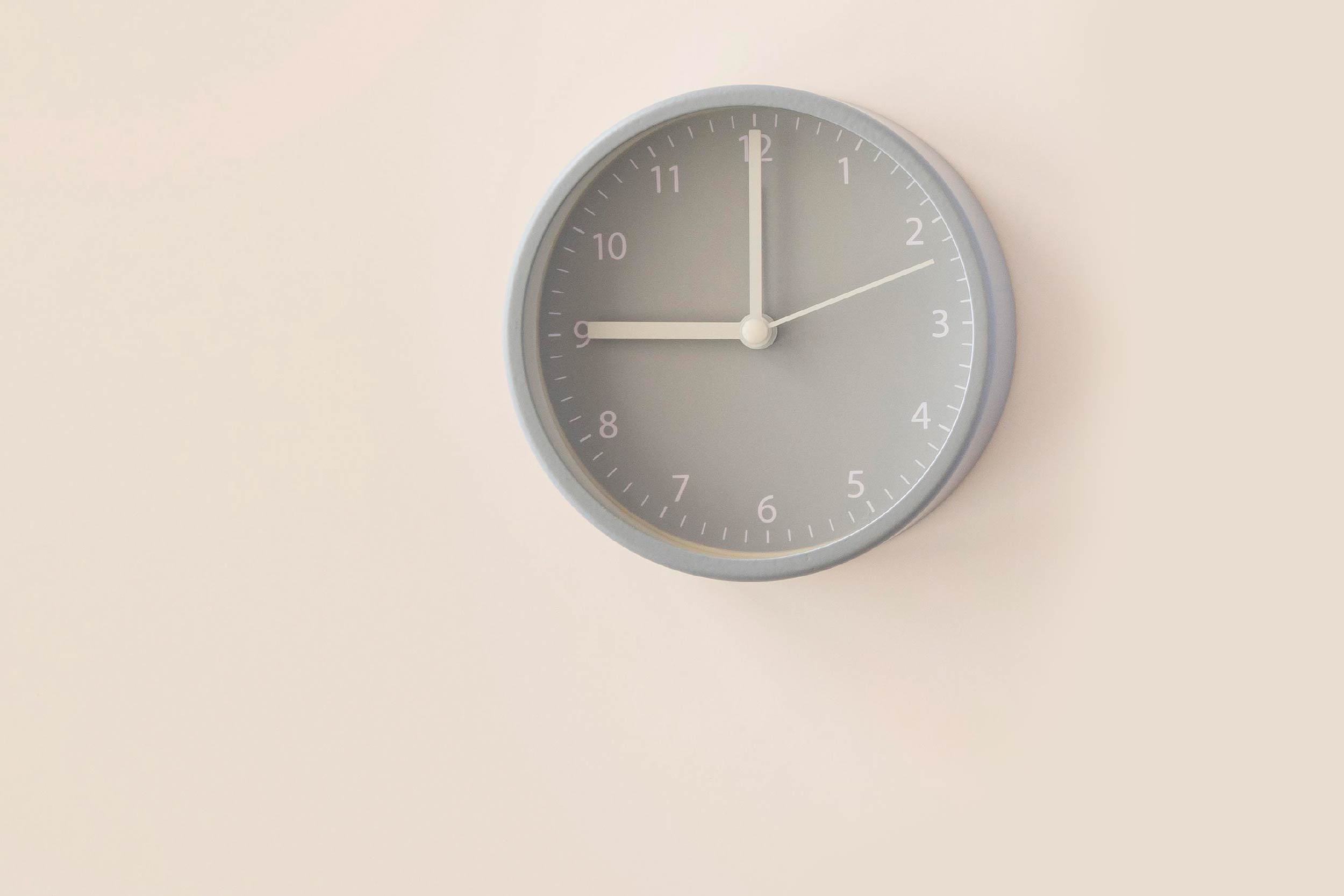 Imagem de um relógio de parede cinza, com ponteiros e numerais brancos. O relógio marca 9 horas e está afixado em uma parede de cor bege.