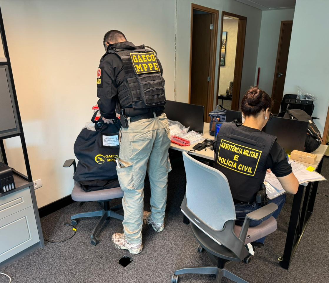 Foto mostra um home e uma mulher, trajados de preto com roupas com a inscrição Gaeco MPPE. Eles estão de frente a uma mesa recolhendo documentos.