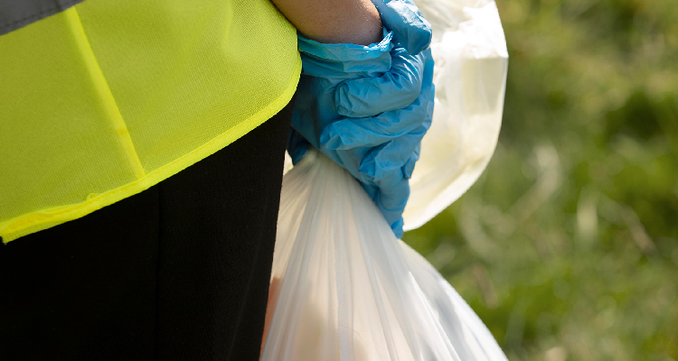 Foto mostra uma mão envolta em luva plástica segurando um saco de lixo branco.