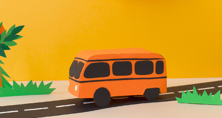 Um ônibus na cor laranja transita da direita para a esquerda sobre uma estrada. Os elementos da imagem são objetos de papel.