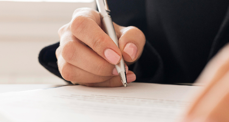 Foto da mão de uma mulher segurando uma caneta prateada. Ela assina um papel.