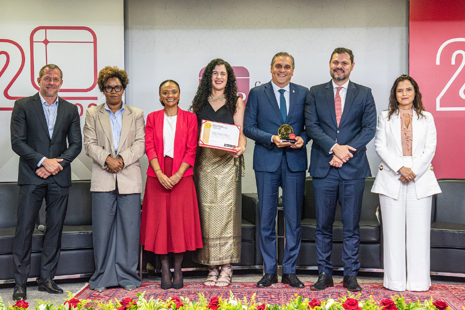 Quatro mulheres e quatro homens, vestindo trajes sociais, posam para uma foto em um evento. A mulher no centro segura um certificado e o homem ao lado segura um prêmio.