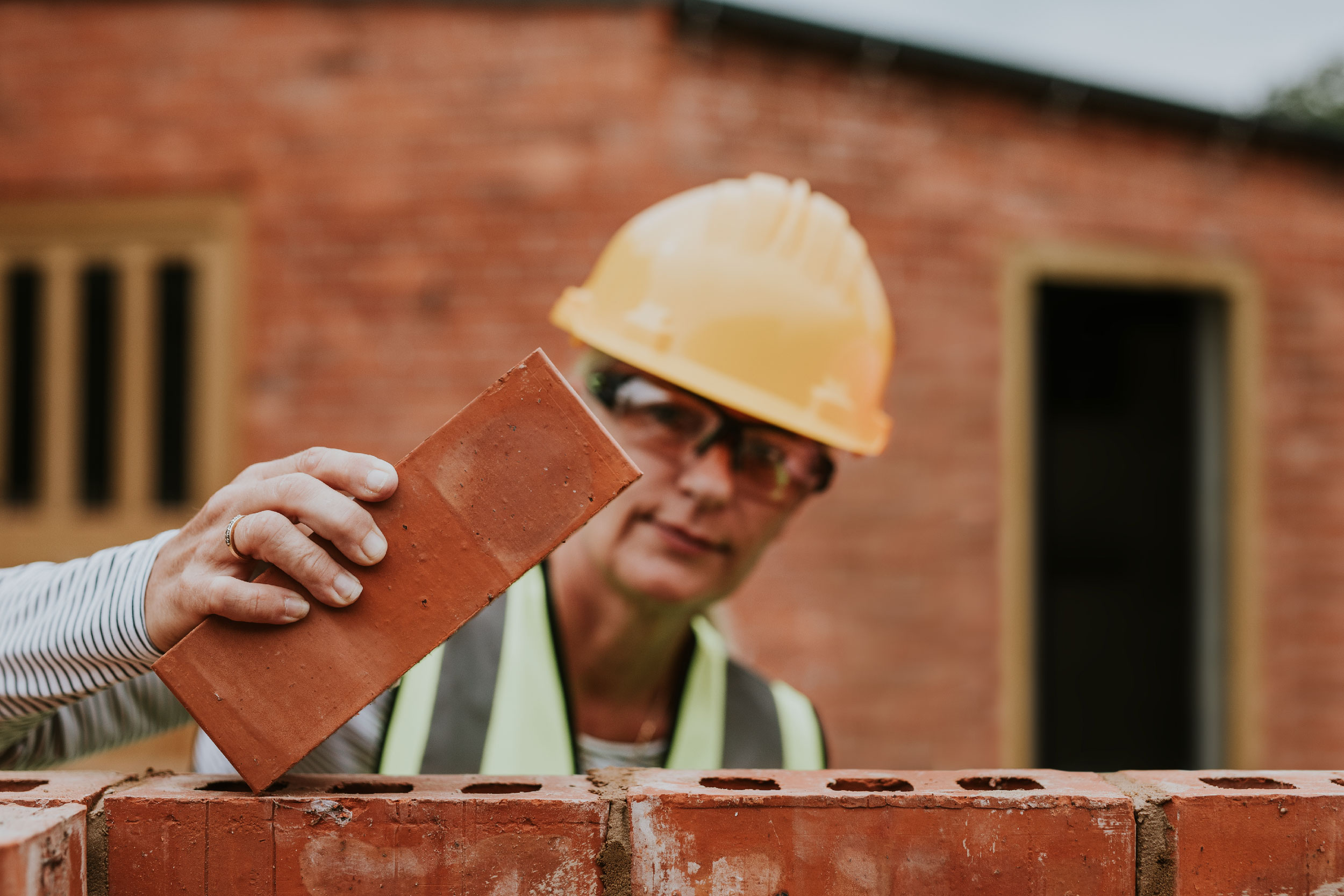 Foto mostra uma pessoa usando capacete com um tijolo na mão. Ela está atrás de uma parede em construção.
