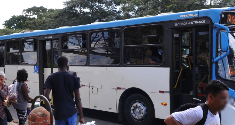 Foto mostra a lateral de um ônibus de cores branca na parte inferior e azul claro na parte superior. Alguns passageiros aguardam para subir no veículo.