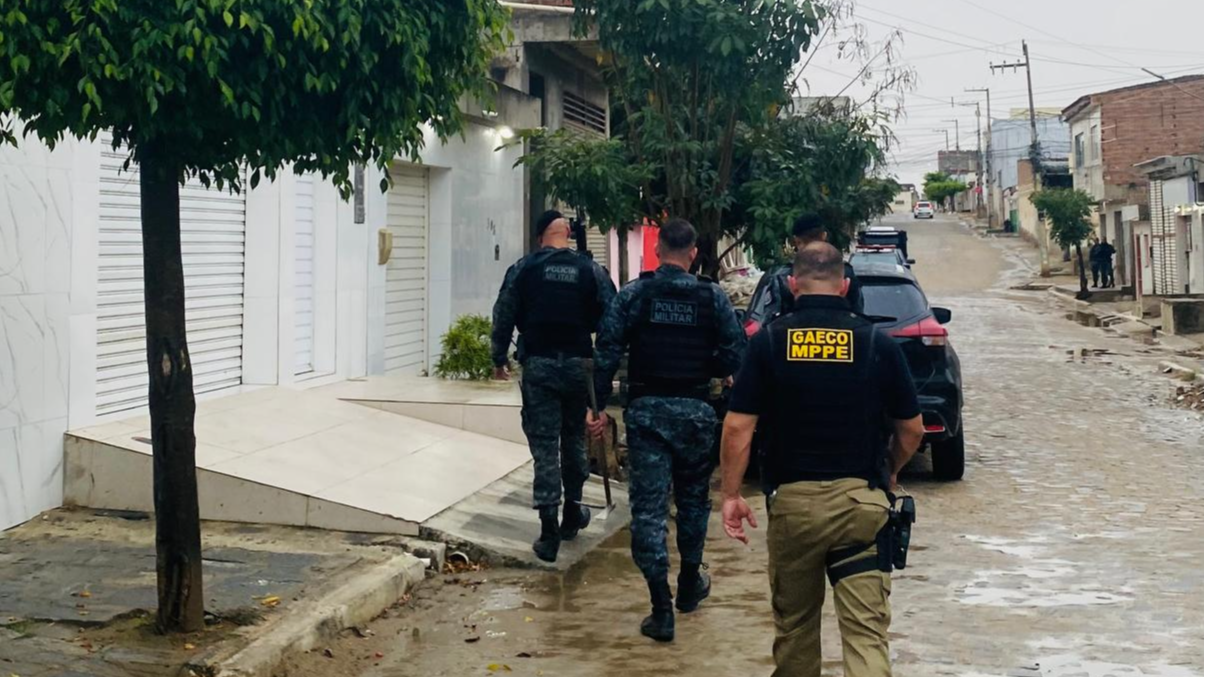 Imagem mostra três homens de costas caminhando por uma rua. Dois deles vestem farda da Polícia Militar e um veste colete do Gaeco MPPE.