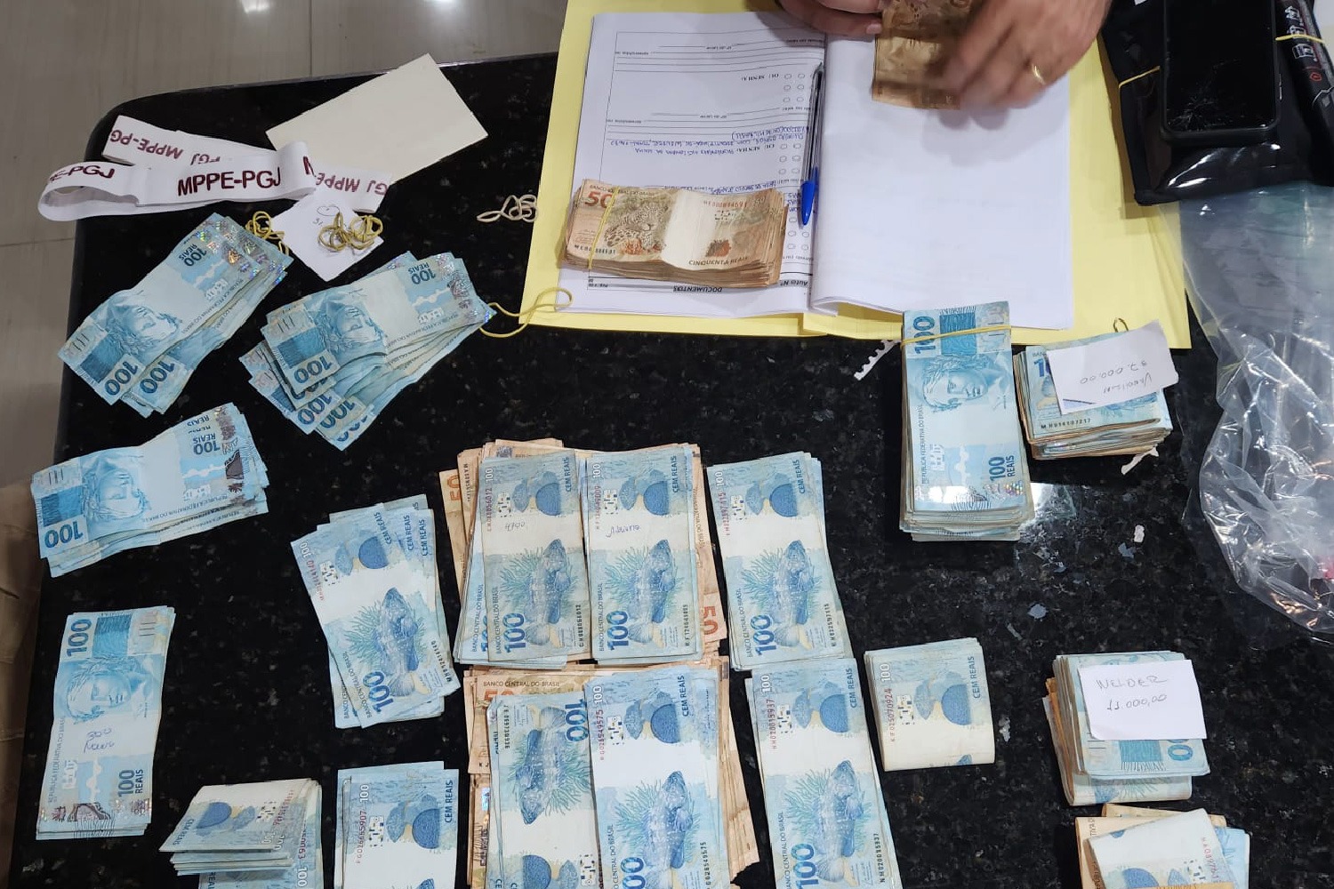 Foto mostra notas de 100 e 50 reais espalhadas sobre mesa de cor preta. No canto superior direito, uma pasta com documentos e uma caneta.