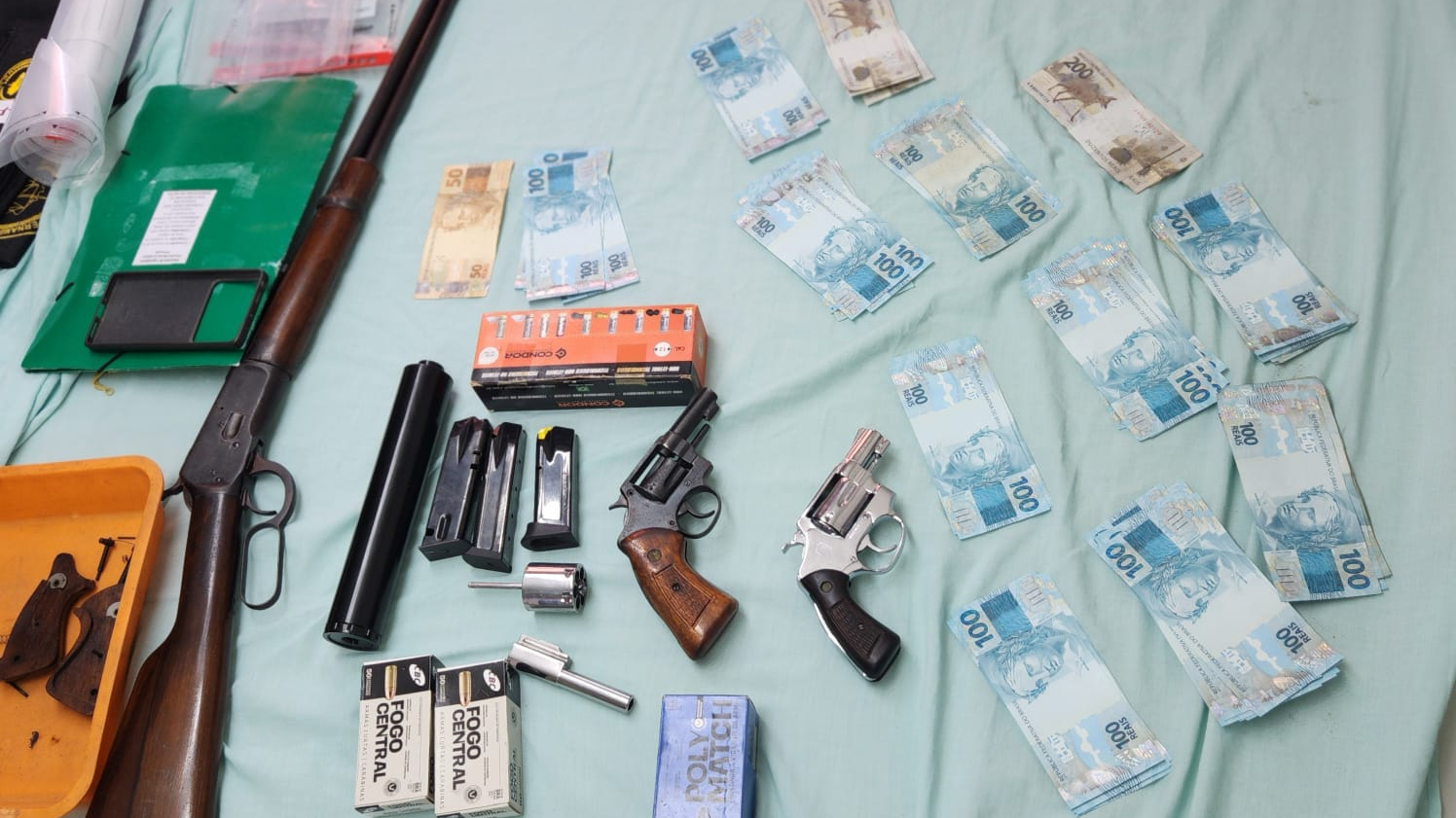 Foto de armas, projéteis e notas de 50 e 100 reais organizados sobre um lençol