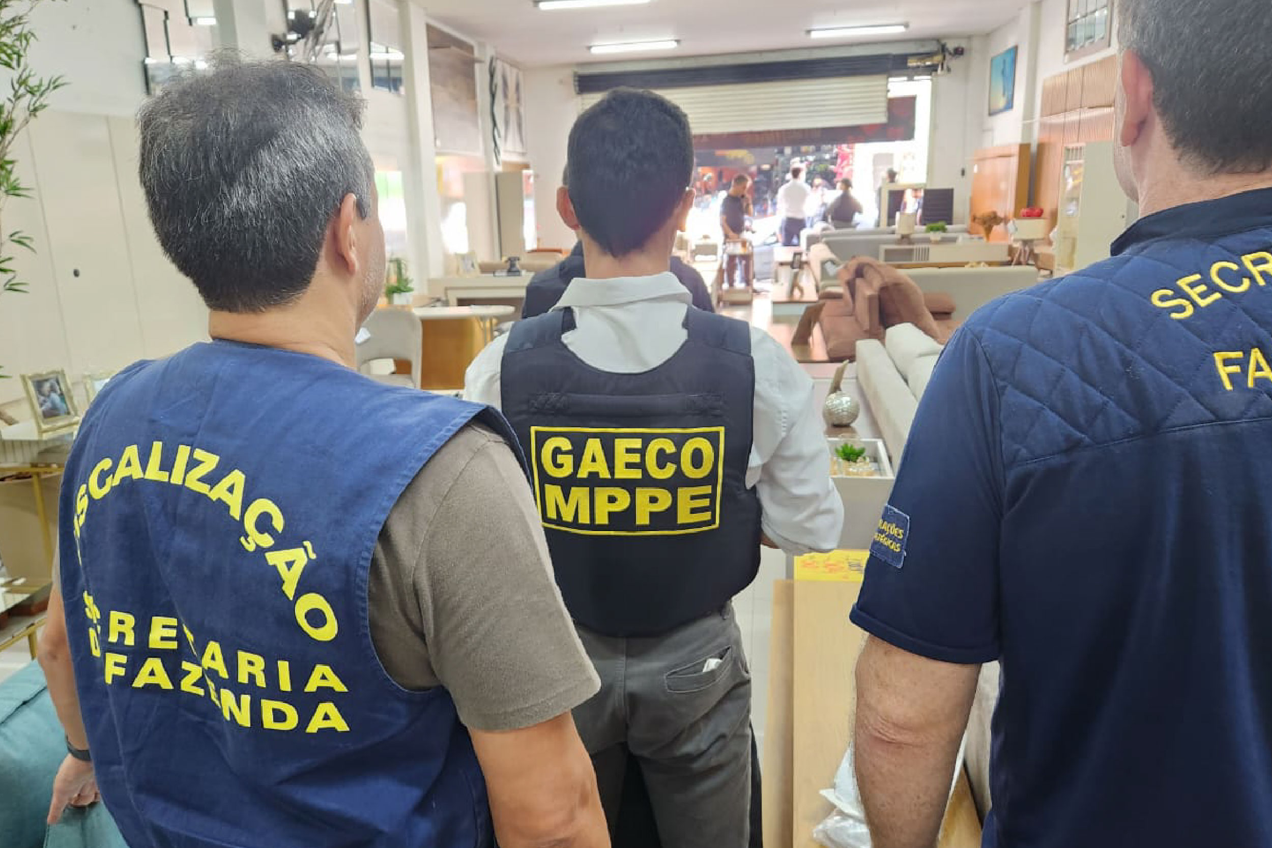 Foto mostra três homens de costas dentro de uma loja. Um deles usa o colete do Gaeco e os outros dois usam coletes da Secretaria da Fazenda.