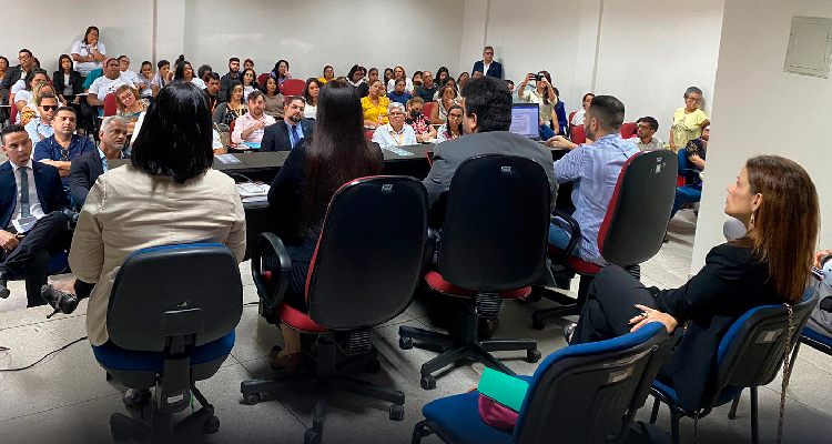Foto mostra três mulheres e dois homens sentados à mesa, de costas para a imagem. Na frente deles, há uma plateia composta por muitas pessoas sentadas em uma sala ampla.