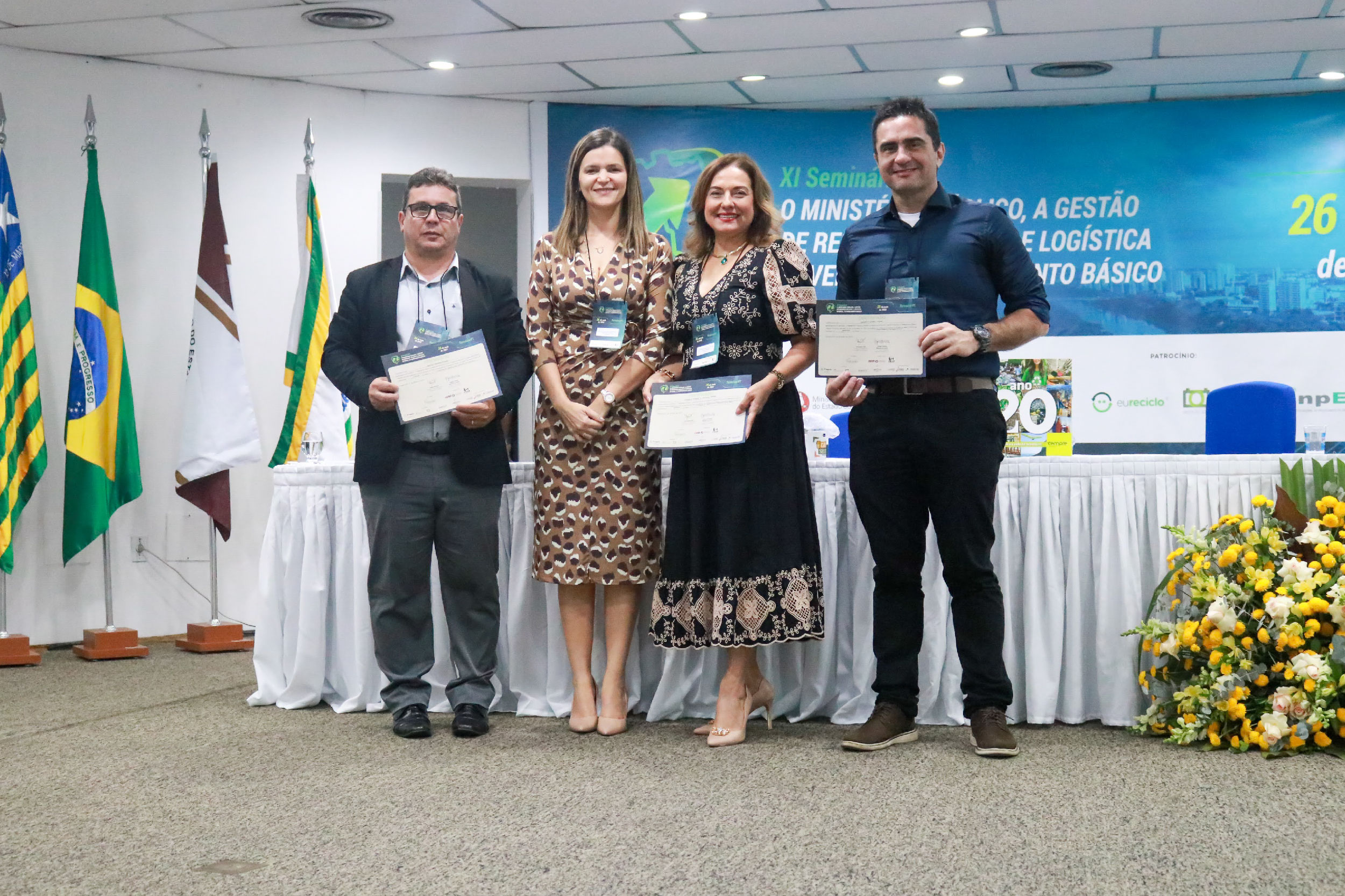 Foto mostra dois homens e duas mulheres de pé, em frente a uma mesa segurando certificados. No canto esquerda imagem há bandeiras do Brasil e do Piauí. No fundo, um banner do evento na cor azul.