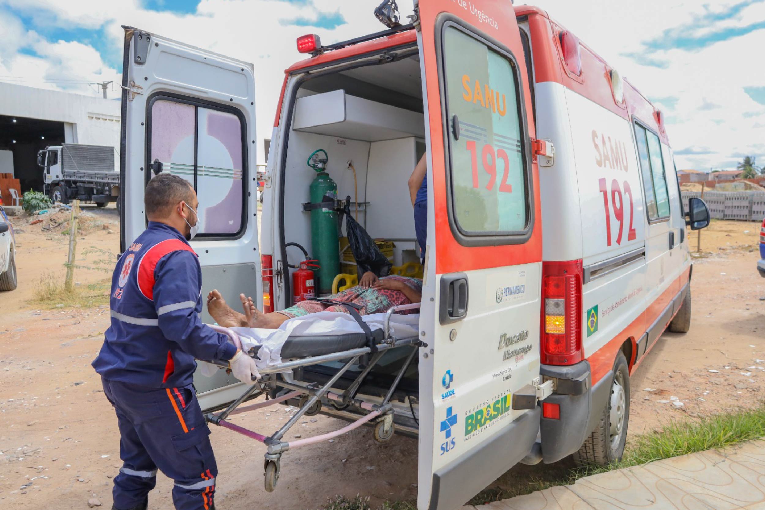 Foto mostra um socorista do SAMU transportando uma pessoa em uma maca para dentro de uma ambulância. O socorrista está de costas e a pessoa na maca está parcialmente dentro da ambulância, sendo possível ser somente suas pernas.