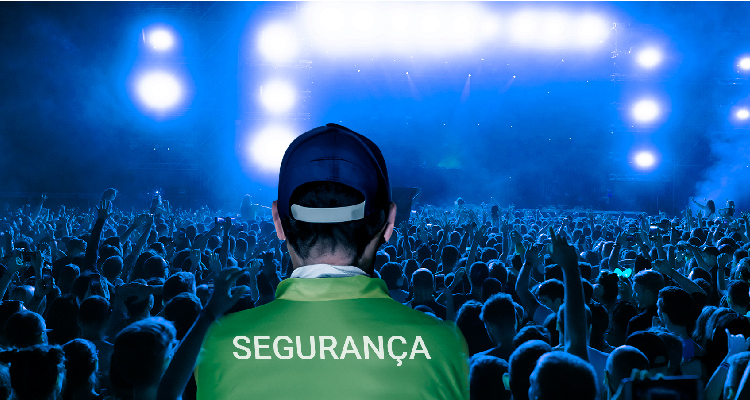 Imagem de homem com colete verde escrito "segurança" olha para multidão que curte show