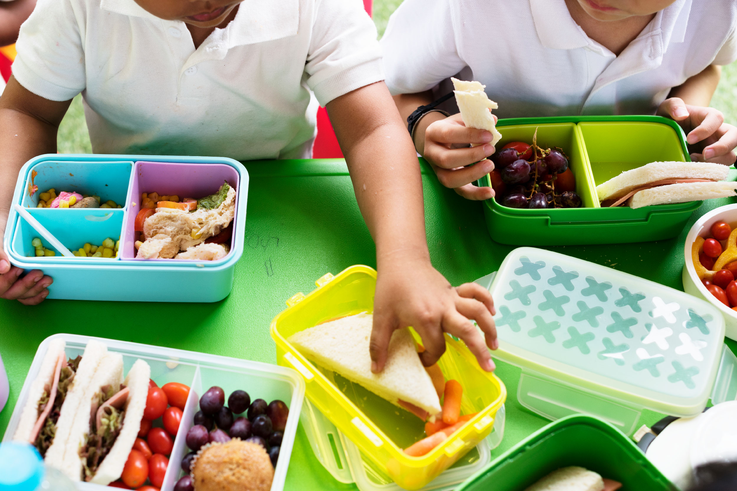 Imagem parcial de crianças comendo lanche sobre uma mesa verde. O lanche está disposto sobre a mesa em potes coloridos e podemos observar frutas, pães e milho.