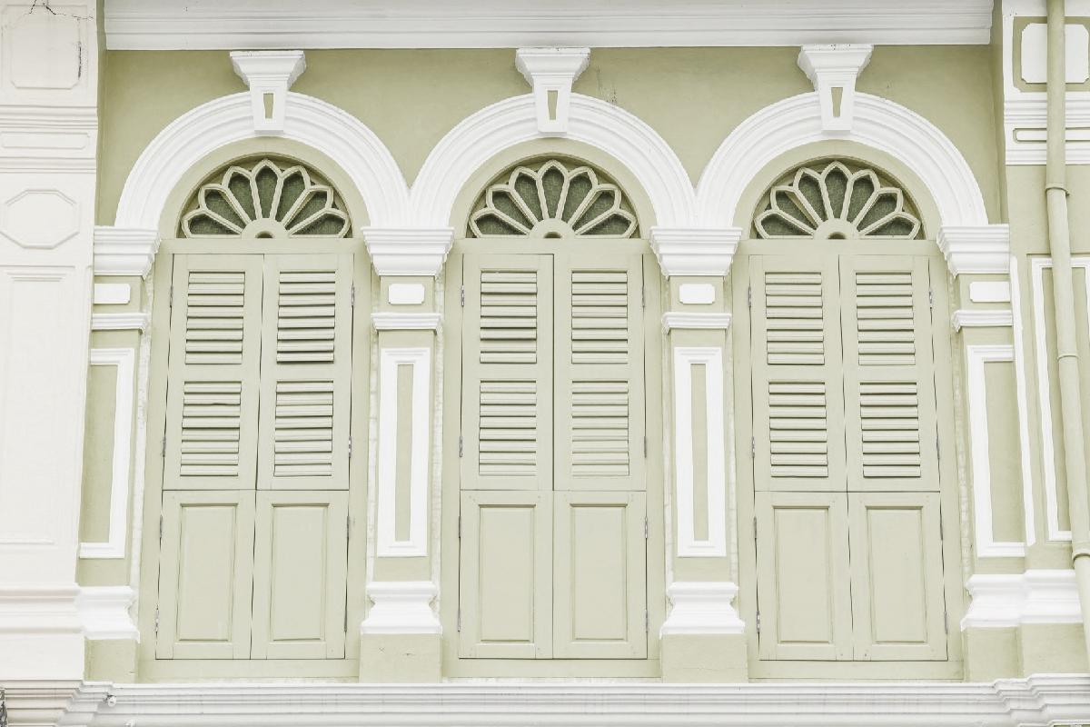 Detalhe da fachada de uma casa, com duas janelas grandes em tons bege, com a parte superior arqueada, com molduras brancas ao redor.