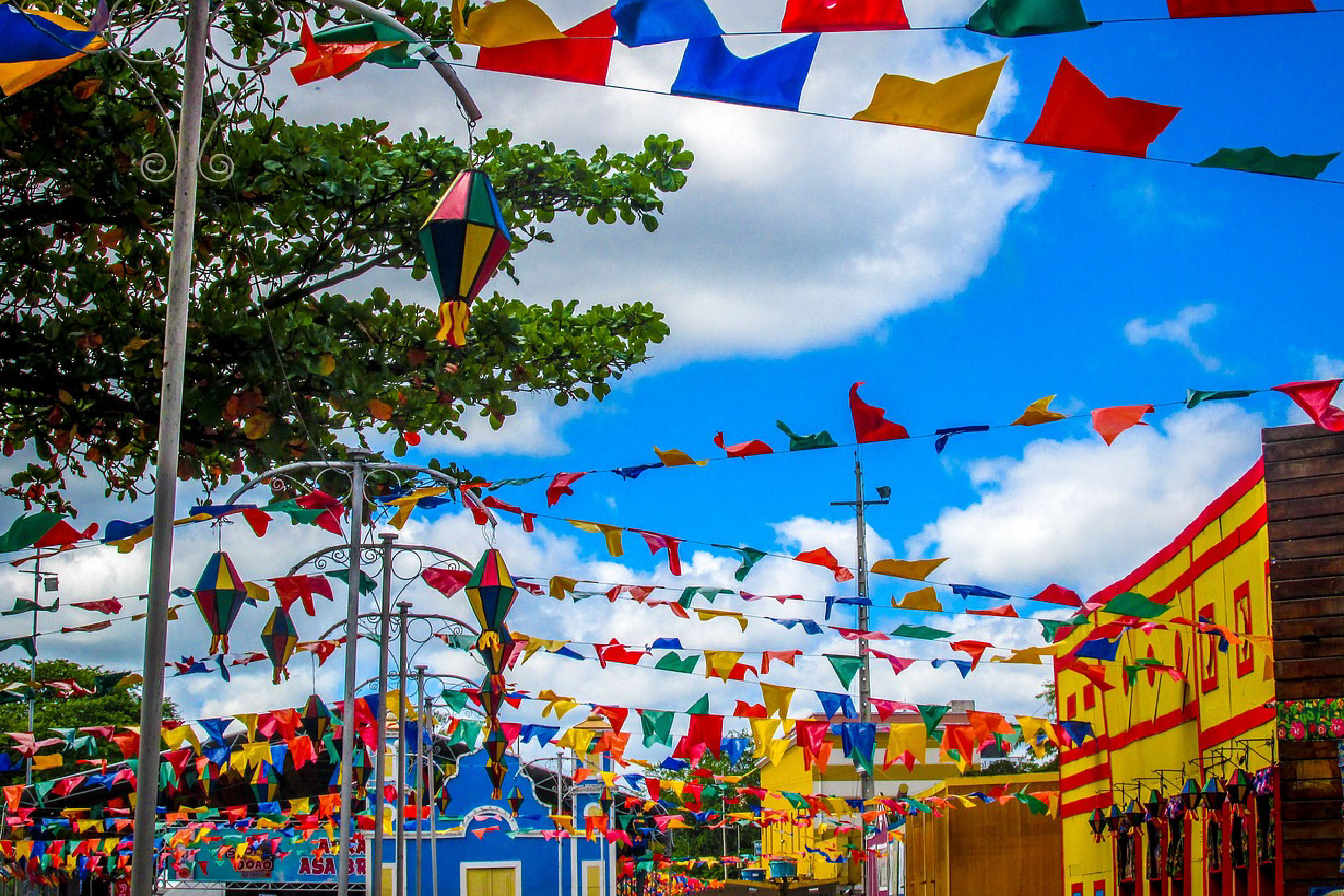 Foto de um conjunto cênico de casinhas coloridas e bandeirolas de São João penduradas na parte superior. No lado esquerdo, alguns postes adornados com balões coloridos.