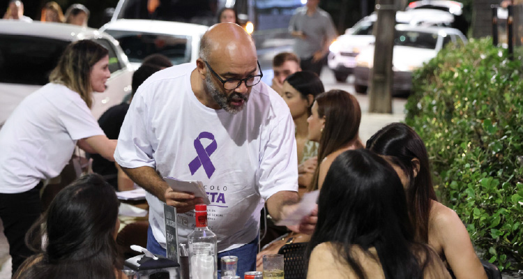 Homem de camisa branca com imagem de laço violeta conversa com duas mulheres que estão sentadas à mesa