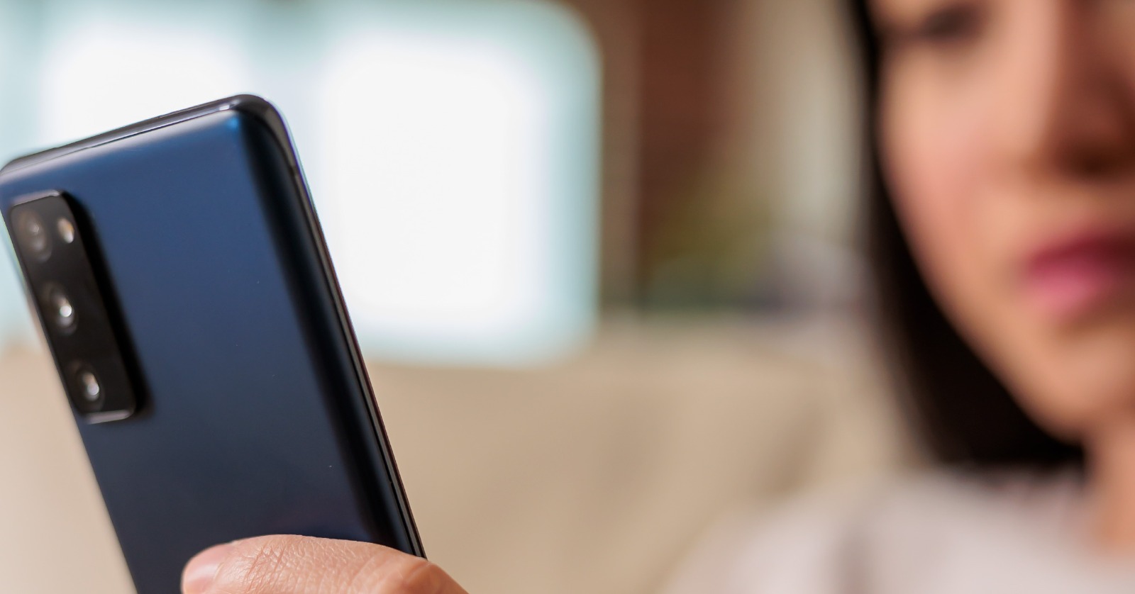 Foto mostra um celular em primeiro plano, do lado esquerdo, segurado por uma mão. No canto direito, em segundo plano, rosto de uma mulher olhando para o aparelho.